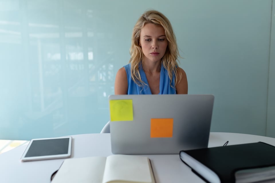 Mujer sentada a un escritorio con un portatil  y varios post-its en la parte posterior de la pantalla. La trabajadora tiene cara de preocupada, como si no fuera capaz de organizar su tiempo de trabajo por sí sola.